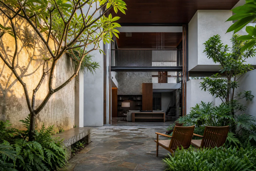 Internal courtyard of The Courtyard House concept — a mature tree and ferns in a planted bed, stone paving, two teak chairs, and the double-height glazed elevation of the house behind. A ModScale warm contemporary villa design concept, Bengaluru.